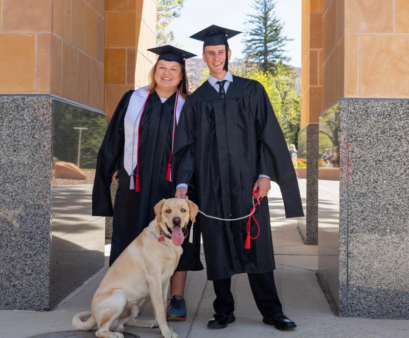 Two graduates from SUU wearing caps and gowns with a dog on a leash