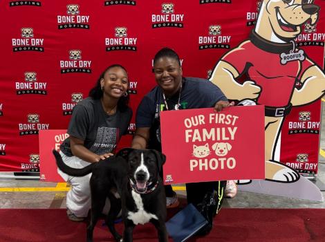 Two people with a dog holding a "Our first family photo" sign in front of a Bone Dry Roofing backdrop with a dog mascot
