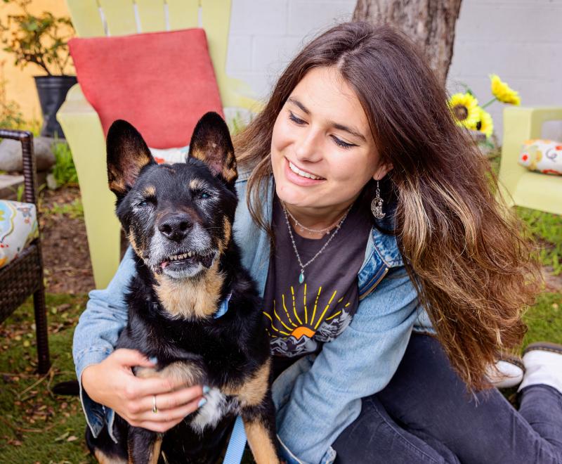 Smiling person sitting on the ground next to a dog while hugging it